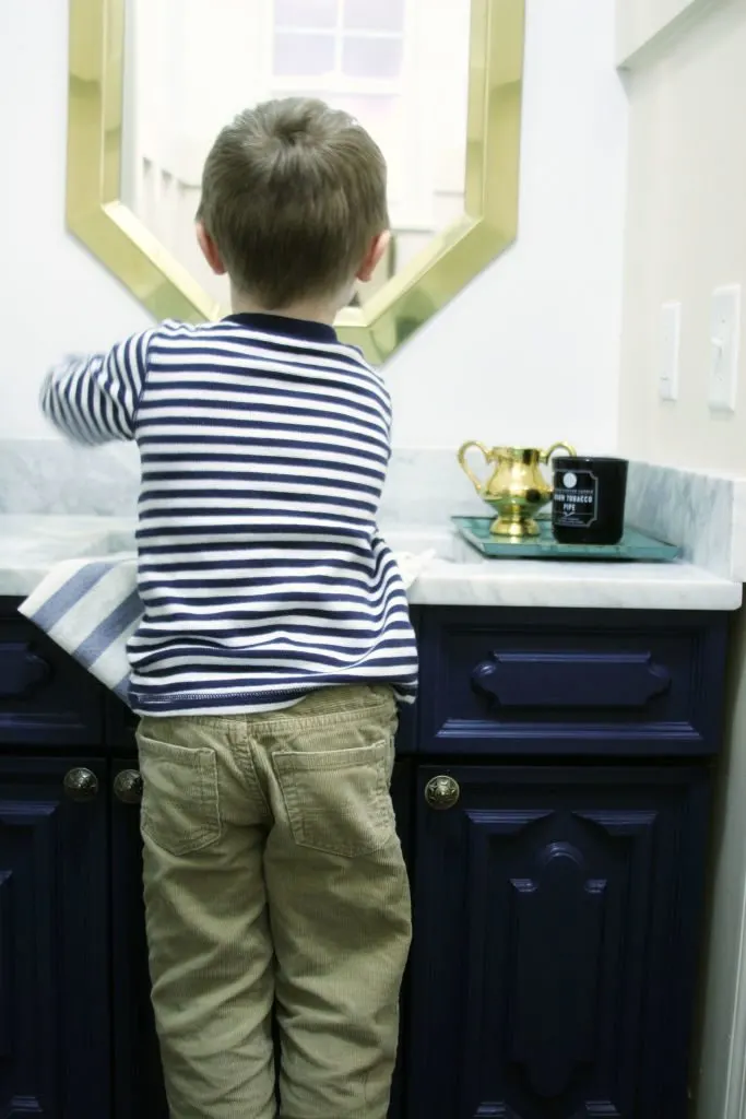 Child washing his hands at a sink with marble counter tops and navy blue cabinets.