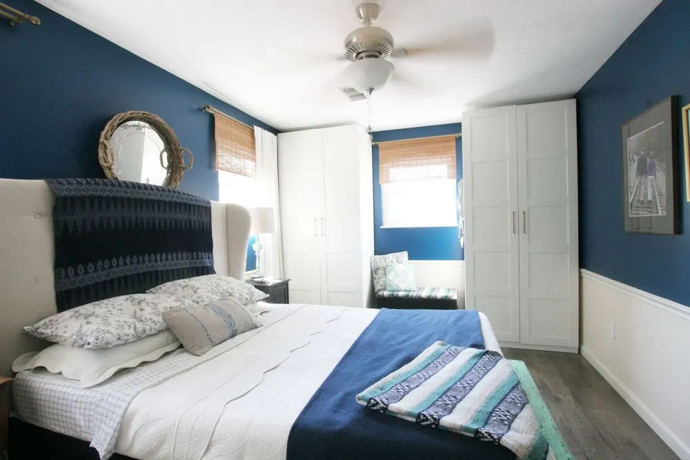 A navy master bedroom, featuring white bedding, bamboo blinds, an upholstered headboard and plenty of storage with large wardrobes and a smart floor plan.