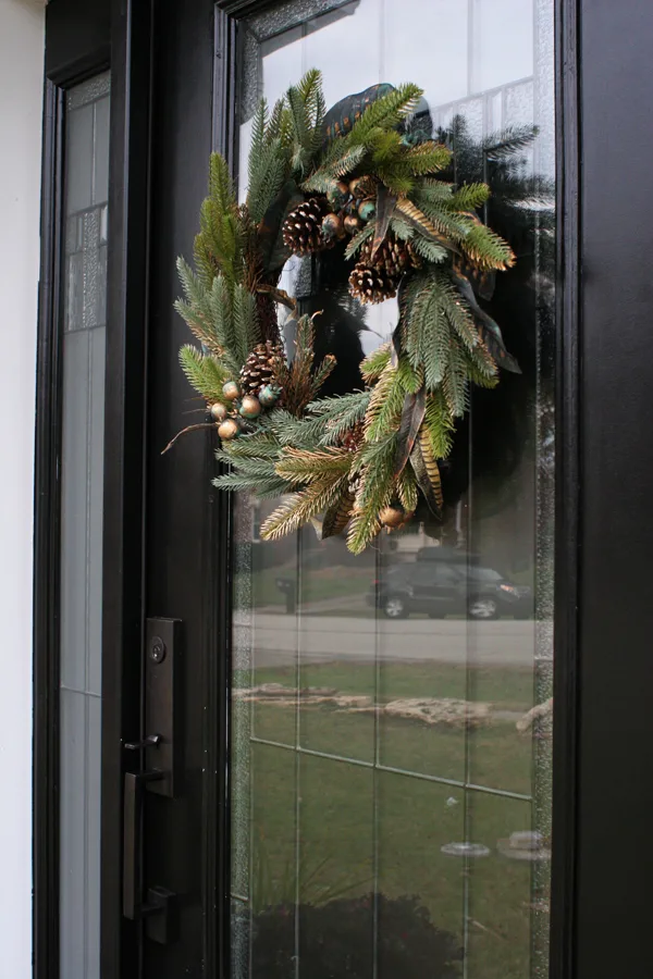 A black front door decorated for Christmas with a simple magnolia, evergreen, and pinecone, wreath