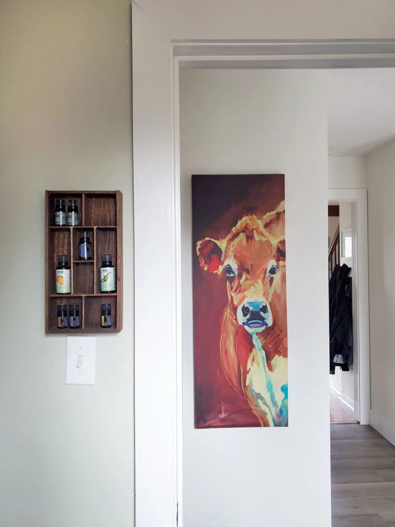 Wooden DIY essential oil shelf hanging on the wall in a laundry room. Cow artwork in hallway. 