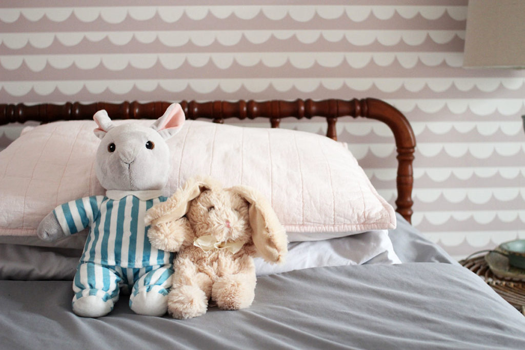 Two stuffed animals on a jenny lind bed with pink and white wallpaper in the background. 