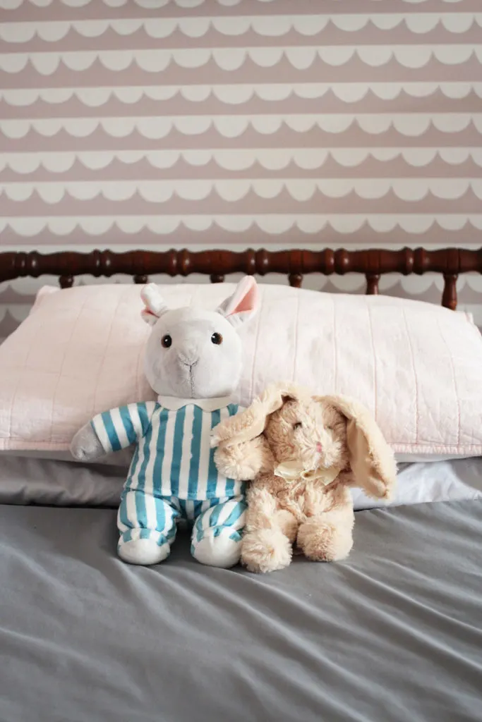 Two stuffed animals on a jenny lind bed with pink and white wallpaper in the background. 